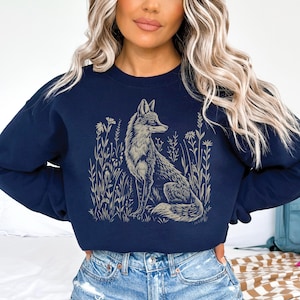 Puede incluir: Una sudadera azul marino con un gr&aacute;fico de un zorro sentado en un campo de flores. El zorro est&aacute; delineado en un color marr&oacute;n claro.