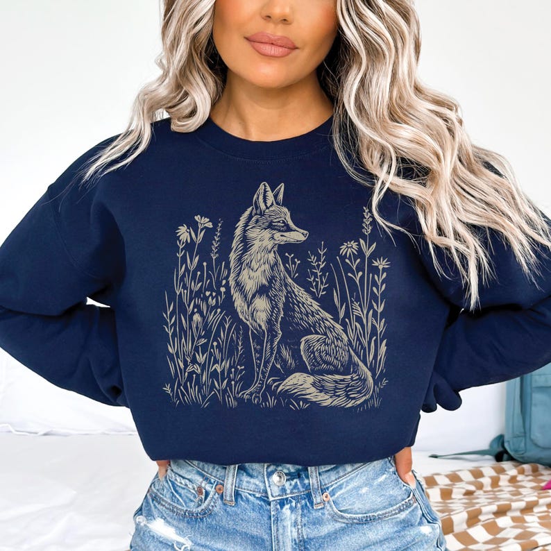 Puede incluir: Una sudadera azul marino con un gr&aacute;fico de un zorro sentado en un campo de flores. El zorro est&aacute; delineado en un color marr&oacute;n claro.