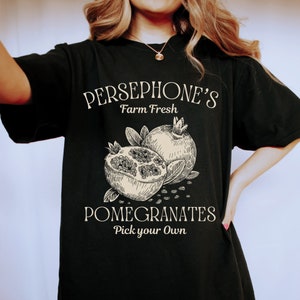 May include: Black t-shirt with a white graphic design that says "Persephone's Farm Fresh Pomegranates Pick your Own". The graphic also features a drawing of two pomegranates.