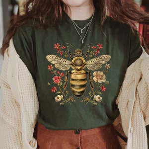 May include: A dark green t-shirt with a vintage illustration of a bee surrounded by flowers. The bee is yellow and black with brown wings. The flowers are red, pink, and white.