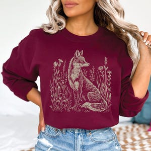 Puede incluir: Una sudadera de manga larga color burdeos con un estampado gr&aacute;fico de un zorro sentado en un campo de flores. El zorro est&aacute; contorneado en un color marr&oacute;n claro.