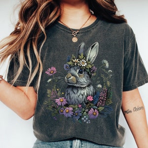 May include: Dark grey t-shirt featuring a detailed illustration of a rabbit wearing a floral crown, surrounded by colorful flowers. The rabbit has grey fur and is set against a backdrop of various wildflowers. The shirt is a comfortable, casual style.