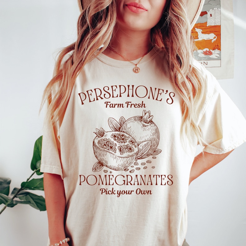 May include: A white t-shirt with brown text that reads "PERSEPHONE'S Farm Fresh POMEGRANATES Pick your Own". The shirt features a drawing of two pomegranates.
