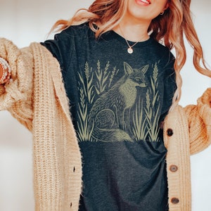 K&ouml;nnte beinhalten: Ein dunkelgraues T-Shirt mit einem Aufdruck eines Fuchses in einem Feld mit hohem Gras. Der Fuchs ist in hellbrauner Farbe umrandet.