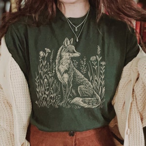 May include: A dark green t-shirt with a cream colored illustration of a fox standing in a field of tall grass. The fox is looking to the right and has a bushy tail. The t-shirt has a crew neck and short sleeves.