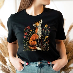 May include: Black t-shirt featuring a vibrant illustration of a fox gazing upwards amidst colorful wildflowers. The fox is depicted in shades of orange, brown, and white, with the flowers in red, yellow, and pink. The design is set against a black background.