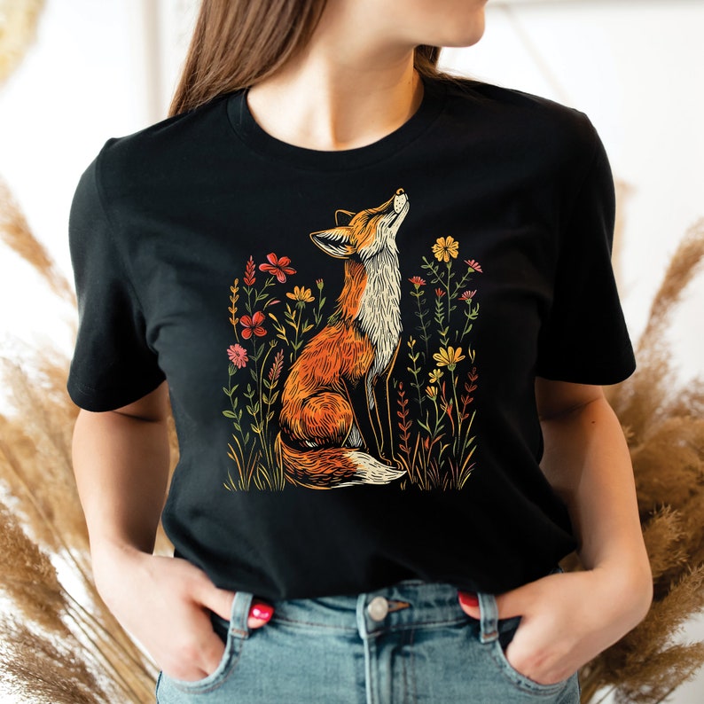 May include: Black t-shirt featuring a vibrant illustration of a fox gazing upwards amidst colorful wildflowers. The fox is depicted in shades of orange, brown, and white, with the flowers in red, yellow, and pink. The design is set against a black background.