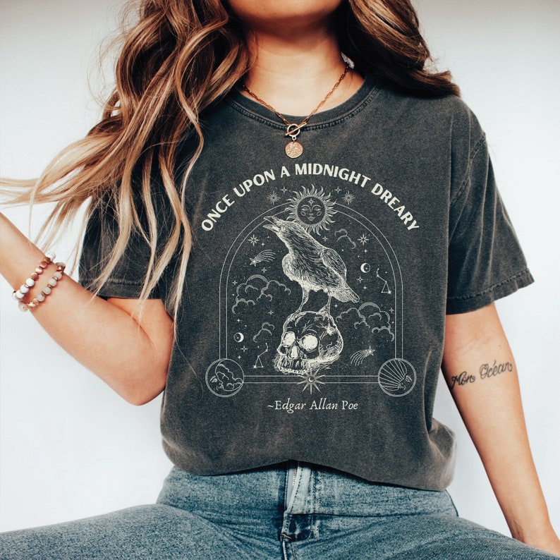 May include: A dark gray t-shirt with a graphic print of a raven perched on a skull. The graphic is surrounded by stars, clouds, and a crescent moon. The text "Once upon a midnight dreary - Edgar Allan Poe" is printed below the graphic.