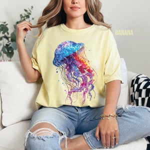 Peut inclure: Une femme portant un t-shirt jaune avec un motif de m&eacute;duse color&eacute;. La m&eacute;duse est une illustration de style aquarelle avec des couleurs bleues, violettes, roses et orange.