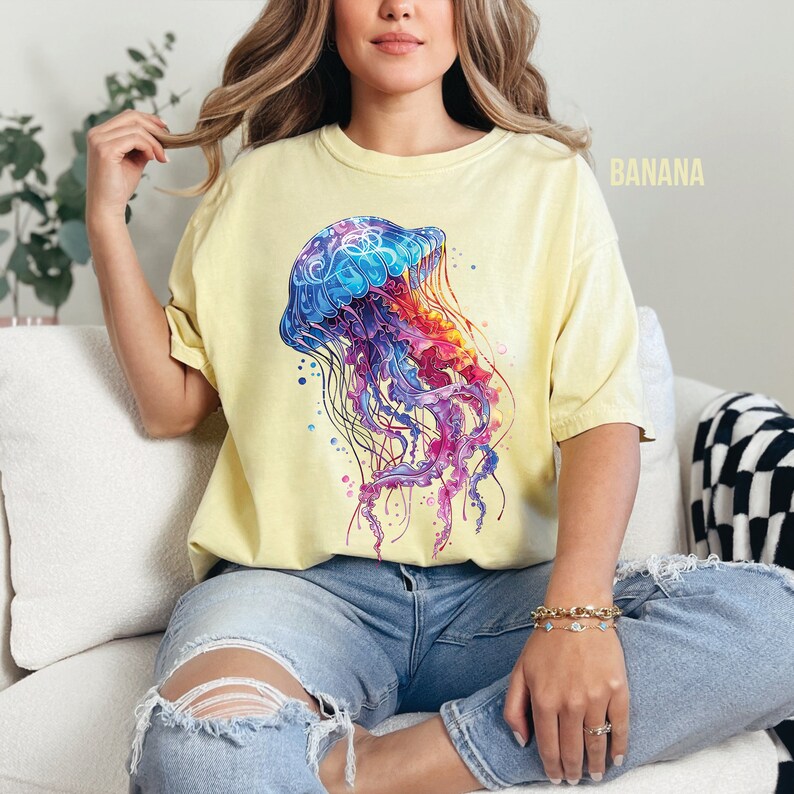 Peut inclure: Une femme portant un t-shirt jaune avec un motif de m&eacute;duse color&eacute;. La m&eacute;duse est une illustration de style aquarelle avec des couleurs bleues, violettes, roses et orange.