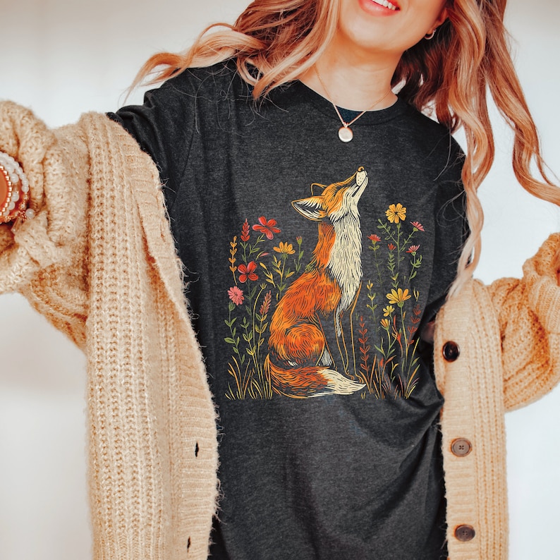 May include: Dark gray long-sleeve shirt featuring a detailed illustration of a fox looking up, surrounded by colorful wildflowers. The fox is orange and white, and the flowers are red, yellow, and pink. A nature-themed graphic tee.
