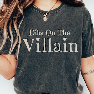 May include: A dark gray t-shirt with the text "Dibs On The Villain" printed in white with two small hearts above and below the word "Villain".