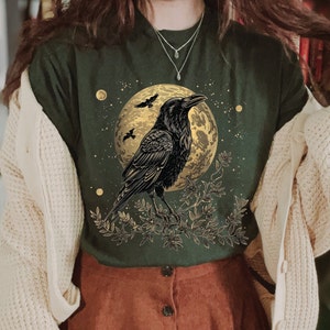 May include: A dark green t-shirt with a black crow perched on a branch in front of a gold moon. The crow is surrounded by small gold stars and the branch has small gold leaves.