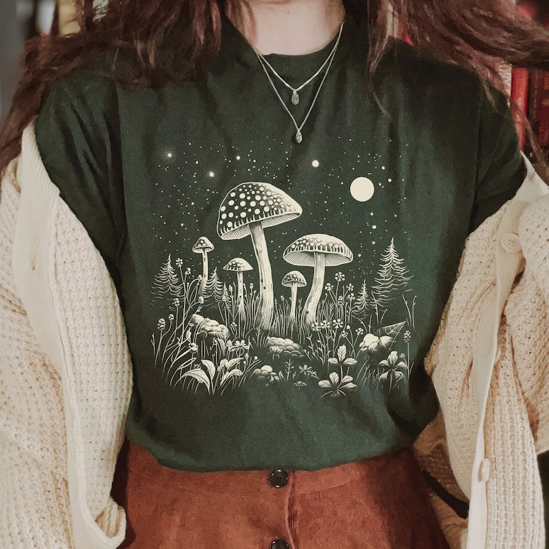 Puede incluir: Camiseta verde oscuro con una ilustraci&oacute;n blanca de setas, plantas, una luna y estrellas. La camiseta se combina con una falda marr&oacute;n y una rebeca color crema. El dise&ntilde;o evoca una escena nocturna del bosque.