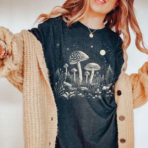Puede incluir: Camiseta de manga larga gris oscuro con una ilustraci&oacute;n crema y beige de setas en un bosque bajo un cielo estrellado. El dise&ntilde;o evoca una est&eacute;tica caprichosa e inspirada en la naturaleza.