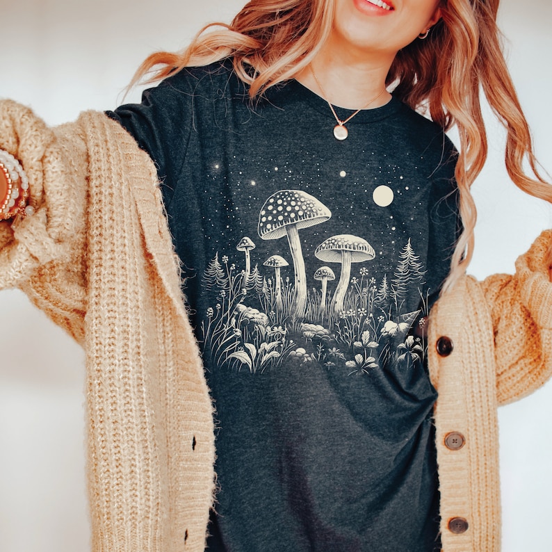 Puede incluir: Camiseta de manga larga gris oscuro con una ilustraci&oacute;n crema y beige de setas en un bosque bajo un cielo estrellado. El dise&ntilde;o evoca una est&eacute;tica caprichosa e inspirada en la naturaleza.
