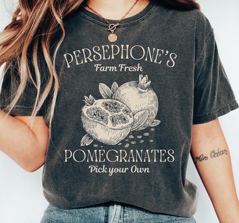 May include: A dark grey t-shirt with a vintage-style graphic design featuring a pomegranate and the text "PERSEPHONE'S Farm Fresh Pomegranates Pick your Own".