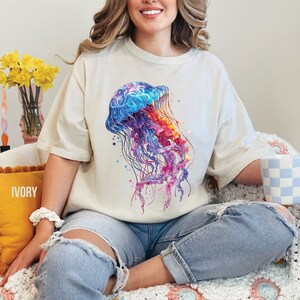Peut inclure: Une femme portant un t-shirt cr&egrave;me avec un motif de m&eacute;duse aquarelle color&eacute;. Le t-shirt est ample et &agrave; manches courtes. La femme est assise sur une couverture en crochet blanche et rose.