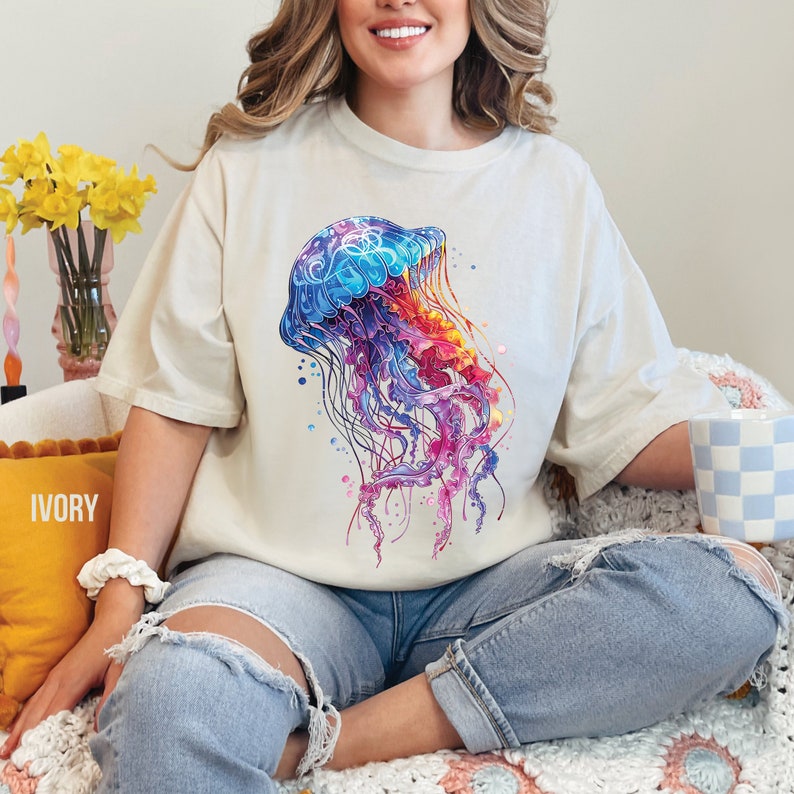 Peut inclure: Une femme portant un t-shirt cr&egrave;me avec un motif de m&eacute;duse aquarelle color&eacute;. Le t-shirt est ample et &agrave; manches courtes. La femme est assise sur une couverture en crochet blanche et rose.