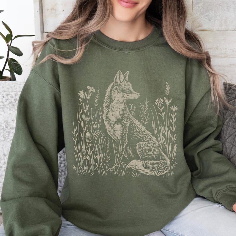 Puede incluir: Una sudadera verde con una ilustraci&oacute;n de color crema de un zorro en un campo de flores. El zorro mira hacia la derecha y tiene una cola tupida.