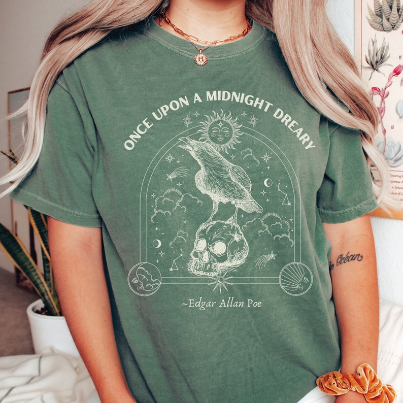 May include: Green t-shirt with a white graphic of a raven perched on a skull with a crescent moon and stars. The text "Once upon a midnight dreary" and "Edgar Allan Poe" are printed on the shirt.