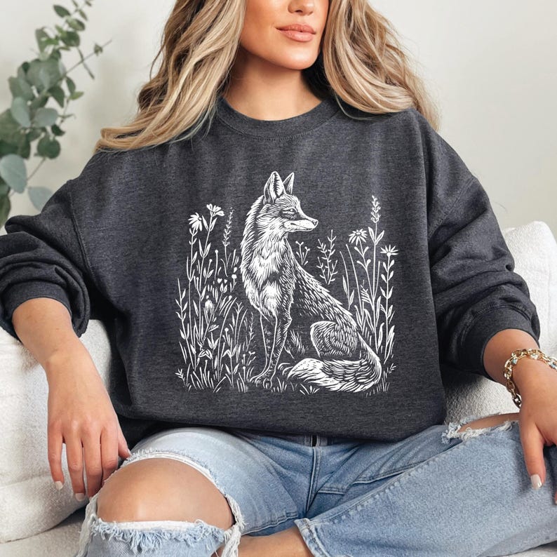 Puede incluir: Una sudadera gris oscuro con un gr&aacute;fico blanco de un zorro sentado en un campo de flores. El zorro mira hacia la derecha y tiene una cola tupida.