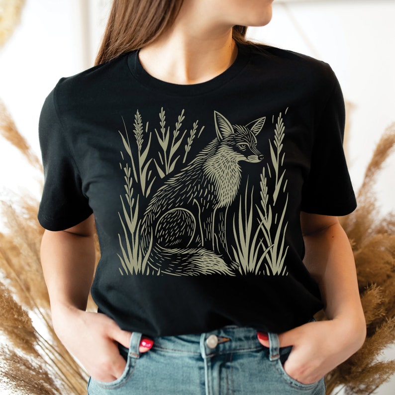 K&ouml;nnte beinhalten: Ein schwarzes T-Shirt mit einem grafischen Aufdruck eines Fuchses, der in hohem Gras sitzt. Der Fuchs ist in hellgr&uuml;n umrandet.