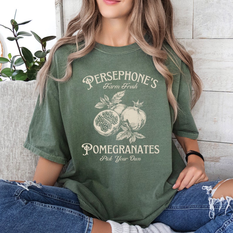 May include: A green t-shirt with a vintage-style graphic design featuring the text "Persephone's Farm Fresh Pomegranates Pick Your Own". The graphic also includes a drawing of pomegranates.