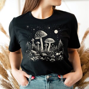 Puede incluir: Camiseta negra con una ilustraci&oacute;n detallada de setas en un entorno forestal bajo un cielo estrellado. El dise&ntilde;o incluye setas de varios tama&ntilde;os, plantas y una luna. El gr&aacute;fico est&aacute; en tonos de blanco y beige.