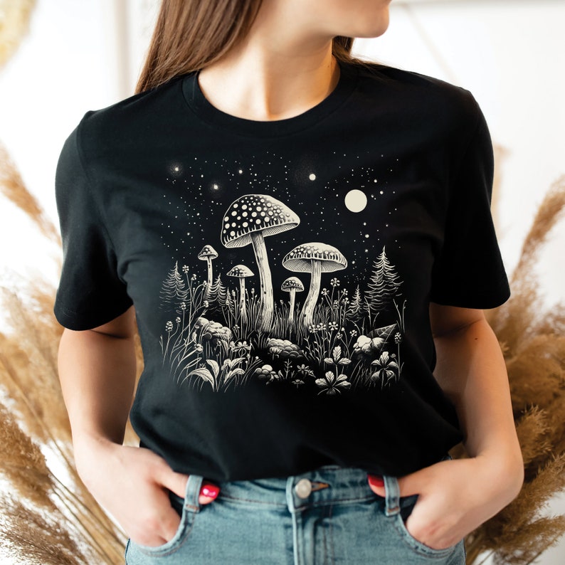 Puede incluir: Camiseta negra con una ilustraci&oacute;n detallada de setas en un entorno forestal bajo un cielo estrellado. El dise&ntilde;o incluye setas de varios tama&ntilde;os, plantas y una luna. El gr&aacute;fico est&aacute; en tonos de blanco y beige.