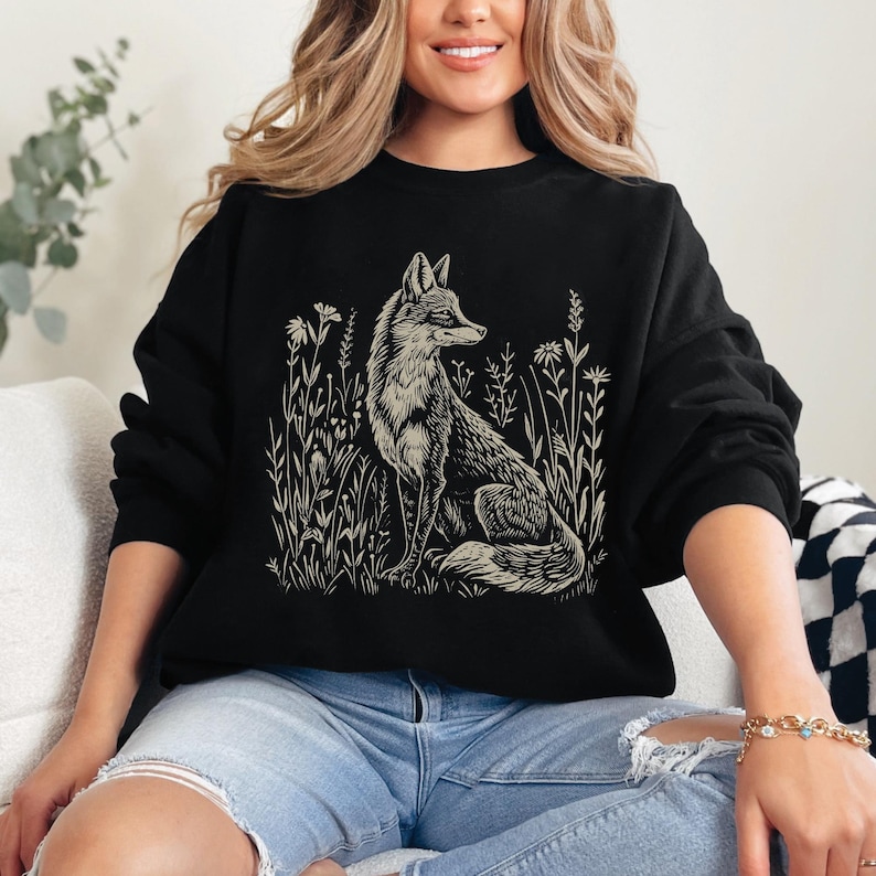 Puede incluir: Sudadera negra con un estampado gr&aacute;fico blanco de un zorro sentado en un campo de hierba alta.