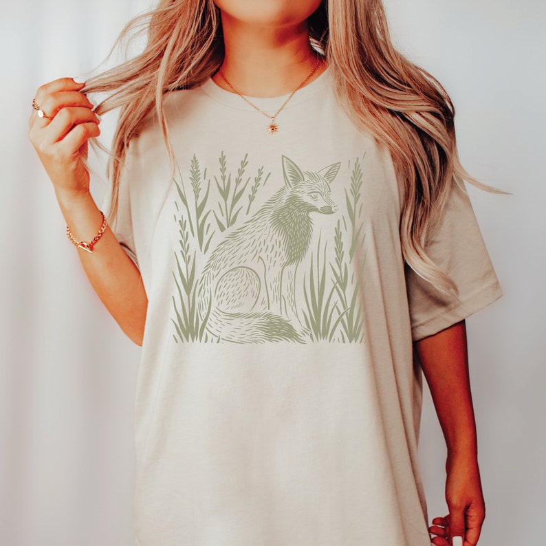 K&ouml;nnte beinhalten: Ein beiges T-Shirt mit einem grafischen Aufdruck eines Fuchses, der in hohem Gras sitzt. Der Fuchs ist in hellgr&uuml;n umrandet.