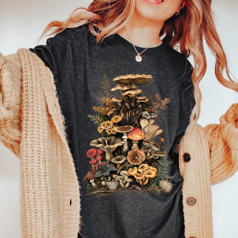 May include: A dark gray t-shirt with a vintage illustration of a cluster of mushrooms in various colors and sizes. The mushrooms are arranged in a tower-like formation, with some mushrooms overlapping others. The illustration is surrounded by leaves and other foliage.