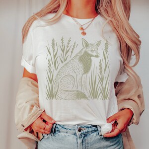 K&ouml;nnte beinhalten: Wei&szlig;es T-Shirt mit einem Aufdruck eines Fuchses, der in hohem Gras sitzt. Der Fuchs ist in hellgr&uuml;n umrandet.