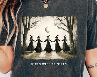 May include: A dark gray t-shirt with a graphic print of five witches in a forest with a crescent moon. The text "Girls Will Be Girls" is printed below the graphic.