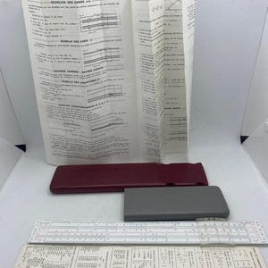 Op de afbeelding: Een vintage rekenliniaal met een rode en grijze koffer en een set instructies. De rekenliniaal is gemaakt van wit plastic en heeft een zwarte schaal. De instructies zijn in het Frans gedrukt en bevatten informatie over het gebruik van de rekenliniaal.
