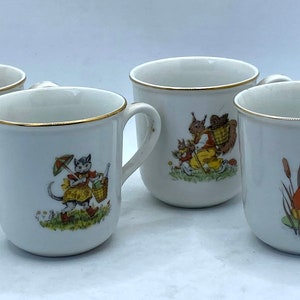 Op de afbeelding: Set van vier witte keramische mokken met gouden rand. Elke mok heeft een andere grillige illustratie van een dier: een varken dat banjo speelt, een kat met een mand, een eekhoorn met een mand en een kikker die trompet speelt.