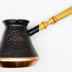 以下が含まれることがあります： 木製のハンドルが付いた黒と銅のトルコ式コーヒーポット。ポットは銅の縁取りが施され、円形のデザインには「アルメニア エレバン」の文字と山脈が描かれています。