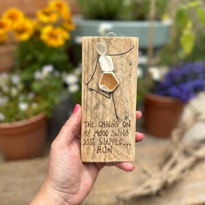 May include: A wooden sign with a white and brown figure made of sea glass. The sign reads "The chains on my mood swing just snapped... Ron".
