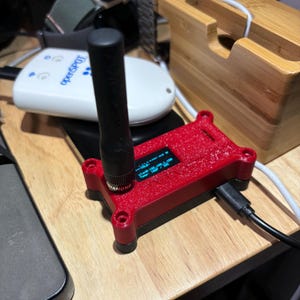 May include: A red 3D printed case with a small screen displaying data and a black antenna. The case is connected to a black cable.