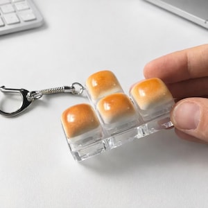 May include: A clear plastic keychain with four square, orange and white, gel-like charms. The charms have a glossy finish and are held in a clear plastic holder. A silver keyring and chain are attached to the holder.
