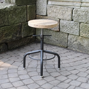 May include: A wooden and metal adjustable height stool with a round seat and a black metal base. The stool is sitting on a gray brick patio.