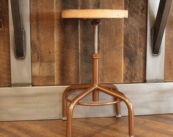 May include: A copper-colored adjustable height stool with a wooden seat. The stool is in front of a rustic wooden wall with metal accents.