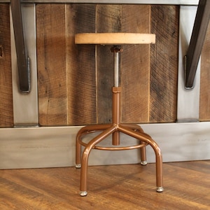 May include: A copper-colored adjustable height stool with a wooden seat. The stool is in front of a rustic wooden wall with metal accents.