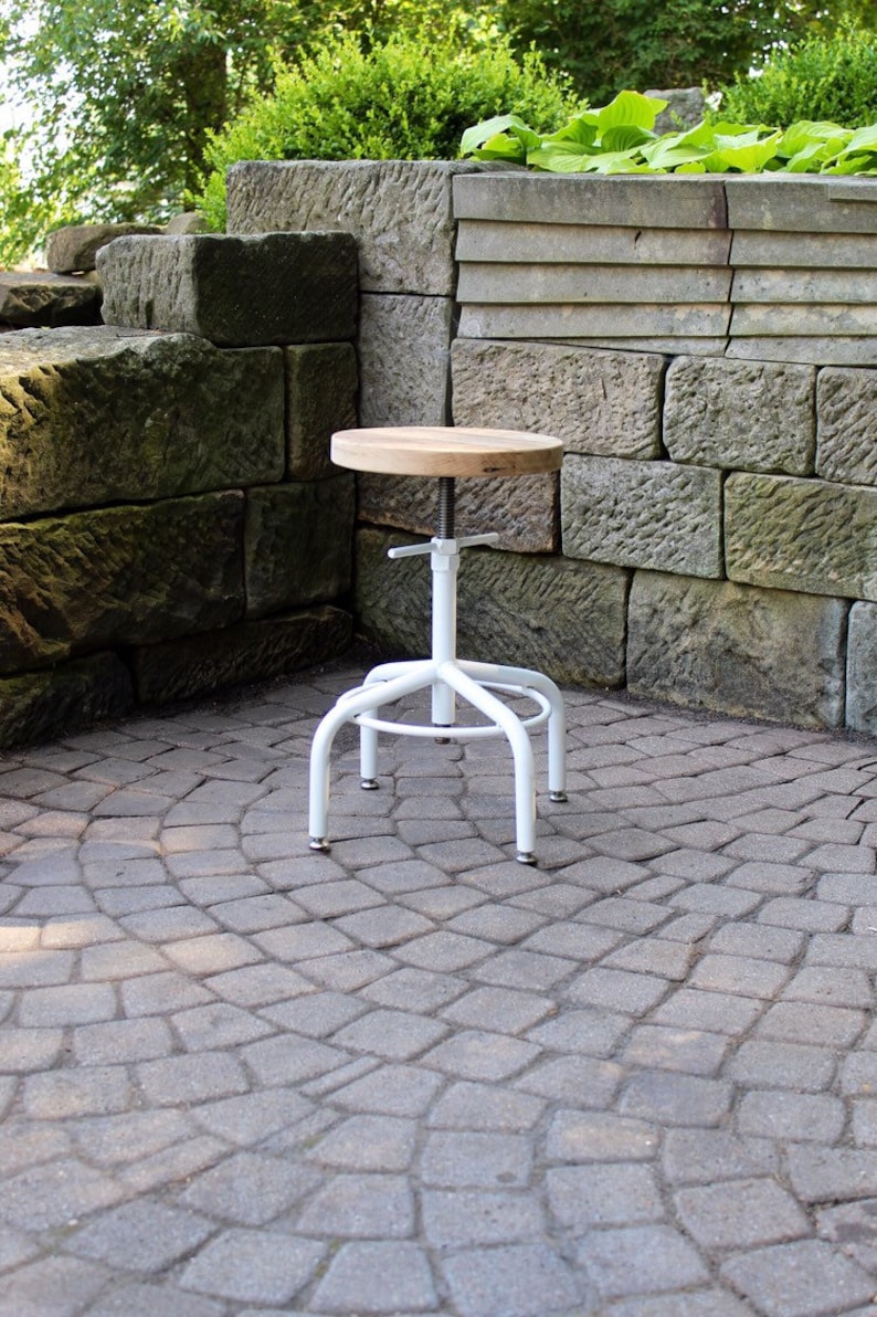 May include: White metal adjustable stool with a round wooden seat. The stool is sitting on a grey stone patio.