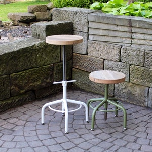 May include: Two industrial style stools with wooden seats. The stool on the left has a white metal base and the stool on the right has a green metal base.