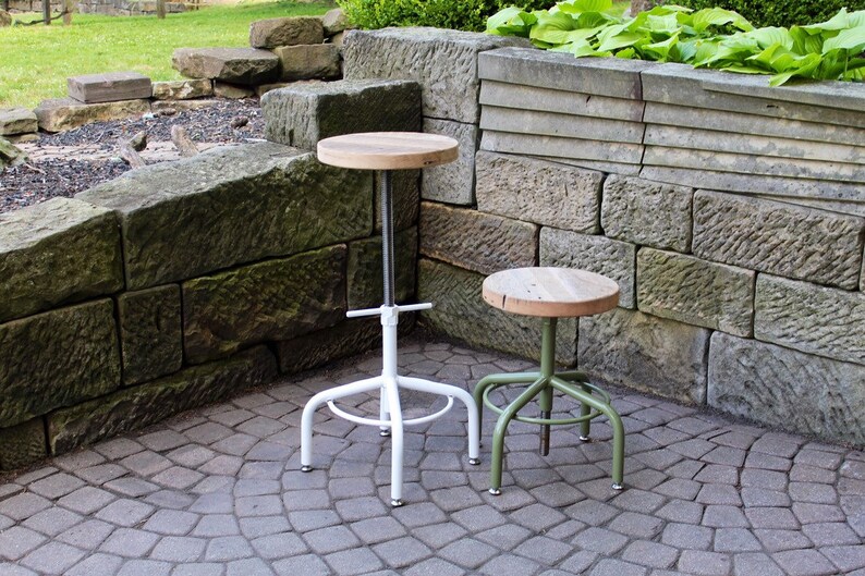 May include: Two industrial style stools with wooden seats. The stool on the left has a white metal base and the stool on the right has a green metal base.