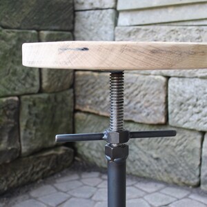 K&ouml;nnte beinhalten: Ein Holzhocker mit runder Sitzfl&auml;che und einem verstellbaren schwarzen Metallfu&szlig;. Der Fu&szlig; hat eine Gewindeschraube und eine Querstrebe zum Einstellen der H&ouml;he.