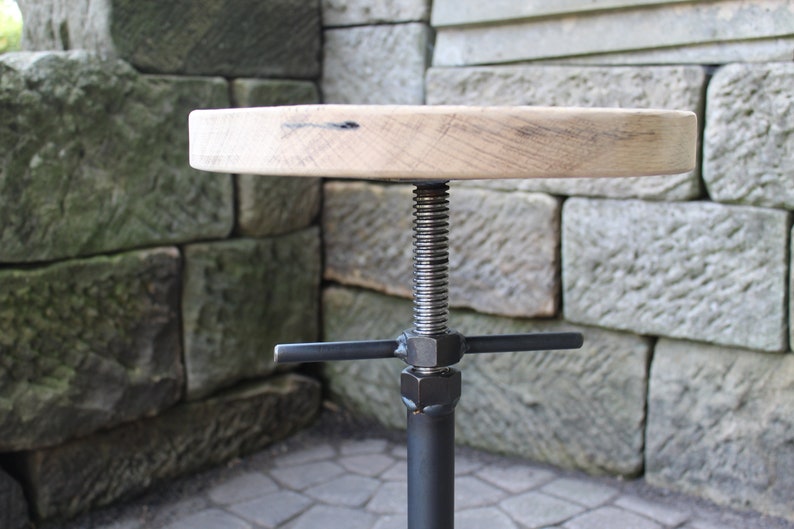 K&ouml;nnte beinhalten: Ein Holzhocker mit runder Sitzfl&auml;che und einem verstellbaren schwarzen Metallfu&szlig;. Der Fu&szlig; hat eine Gewindeschraube und eine Querstrebe zum Einstellen der H&ouml;he.