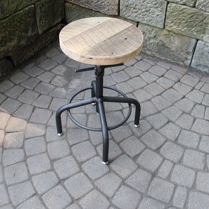 K&ouml;nnte beinhalten: Ein schwarzer Drehhocker aus Metall mit einem runden Holz-Sitz. Der Sitz ist aus recyceltem Holz und hat einen rustikalen Look. Der Hocker steht auf einer grauen Steinplatte.